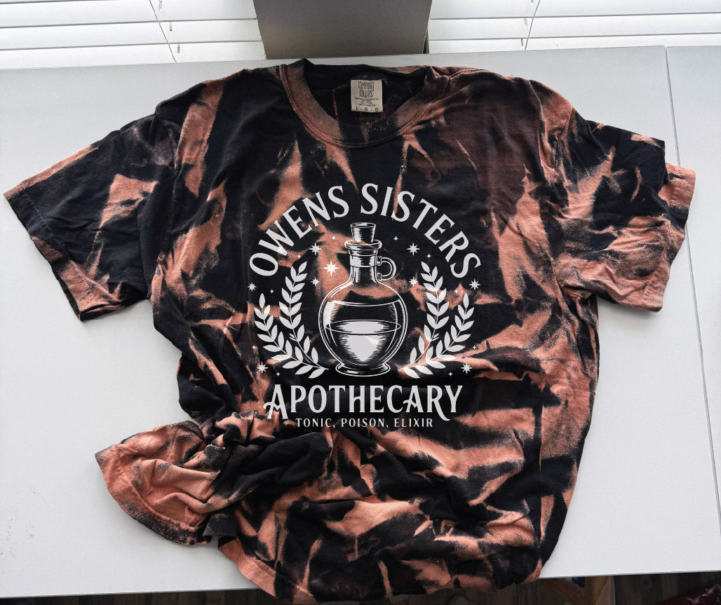 A short-sleeve graphic t-shirt with a black and copper-brown tie-dye pattern, featuring a white printed design of an apothecary's scroll and potion bottle. The text reads 'OWENS SISTERS APOTHECARY' and 'TONIC, POISON, ELIXIR'.